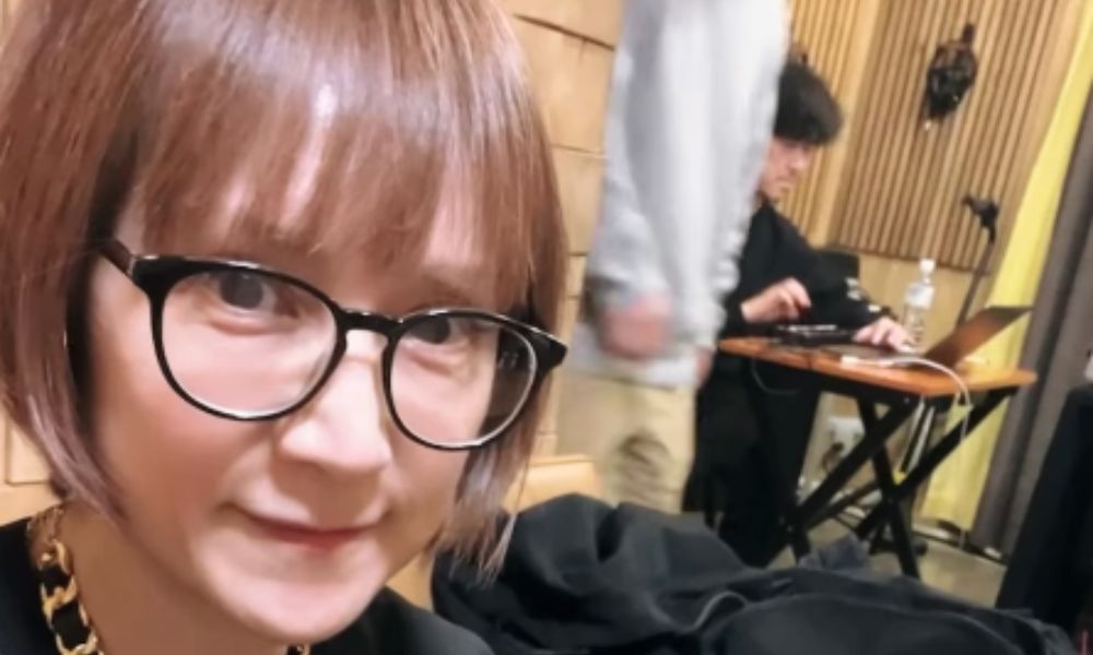 レコーディングスタジオで撮影された鈴木蘭々の自撮り。メガネ姿でカメラを見つめ、後方には機材を操作する音響エンジニアの様子が映っている。