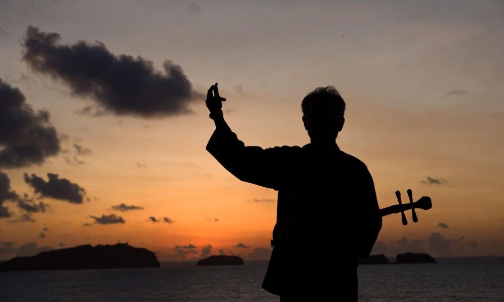夕焼けの海を背景に三味線を持ち、片手を上げる男性のシルエット、兄のイメージを表現した風景