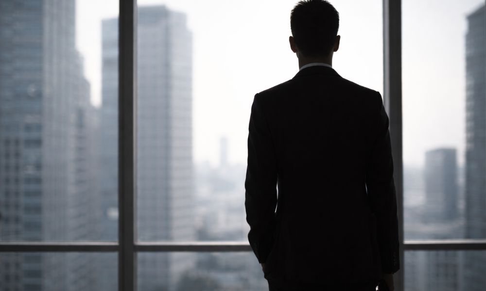 スーツ姿の高身長男性がオフィスの大きな窓の前で街を見下ろす後ろ姿のシルエット